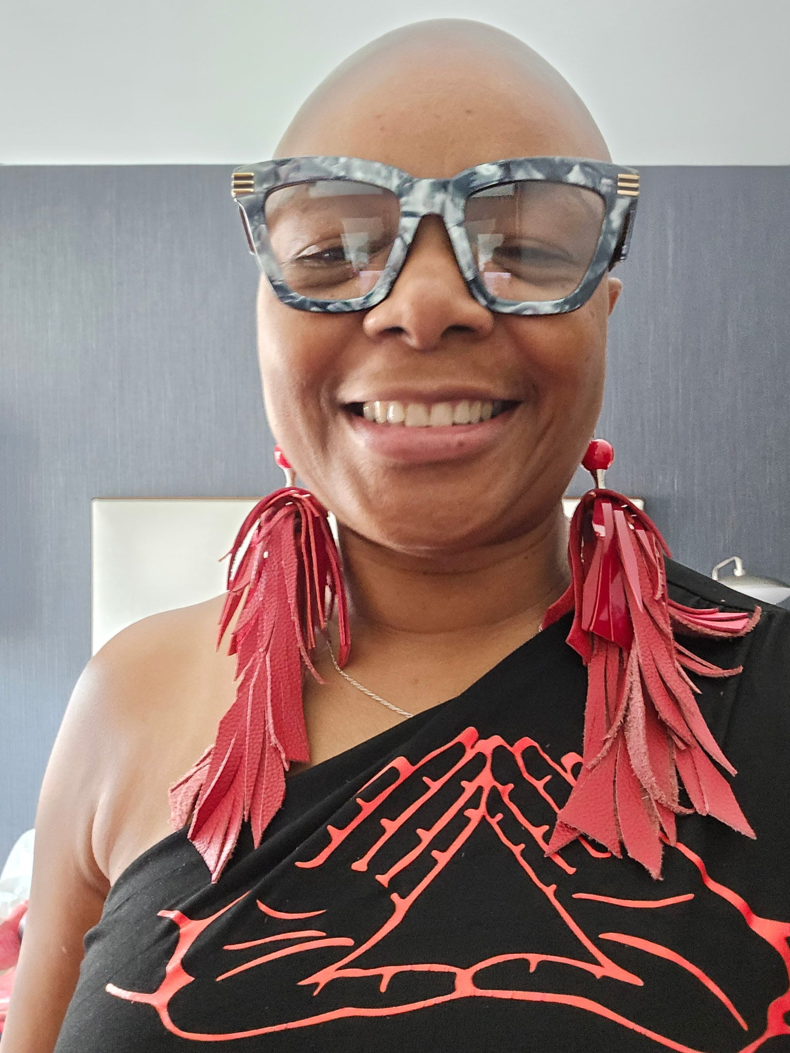 woman wearing large sunglasses and a black top with red mids up designs, sitting against a neutral background.