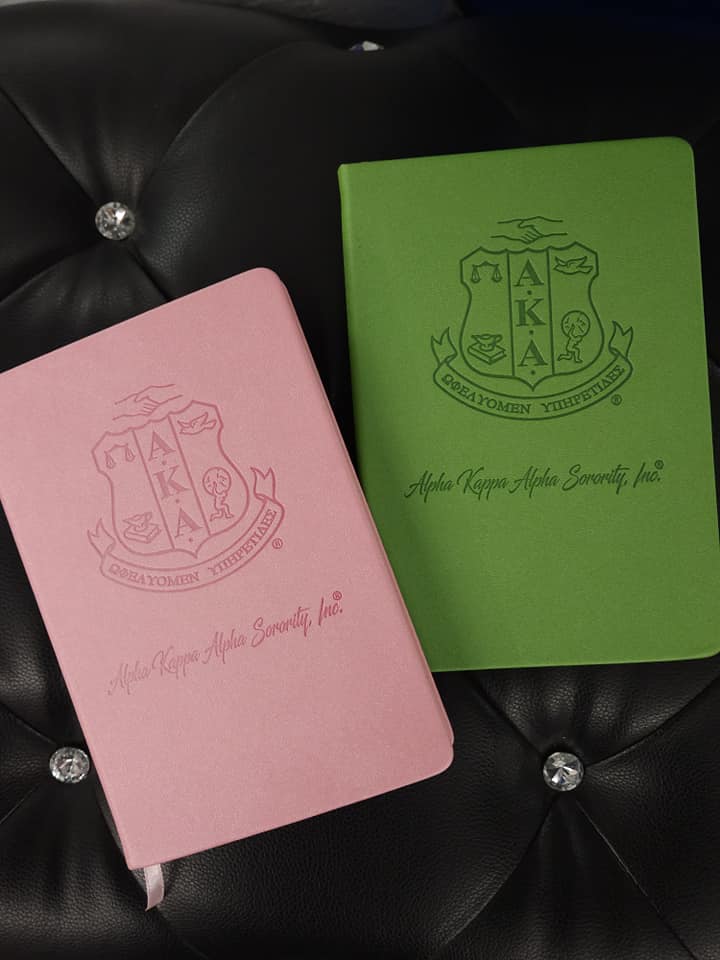 Pink and green notebooks with Alpha Kappa Alpha fraternity logo on a black textured surface.