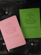 Pink and green notebooks with Alpha Kappa Alpha fraternity logo on a black textured surface.
